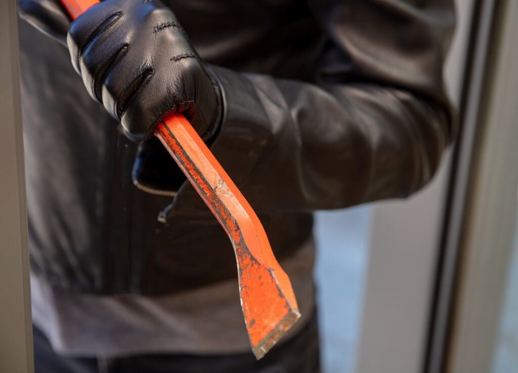 Masked thief entering a house for stealing in the daytime. Burglar in leather jacket breaking a glass aluminum door. Housebreak with crowbar.